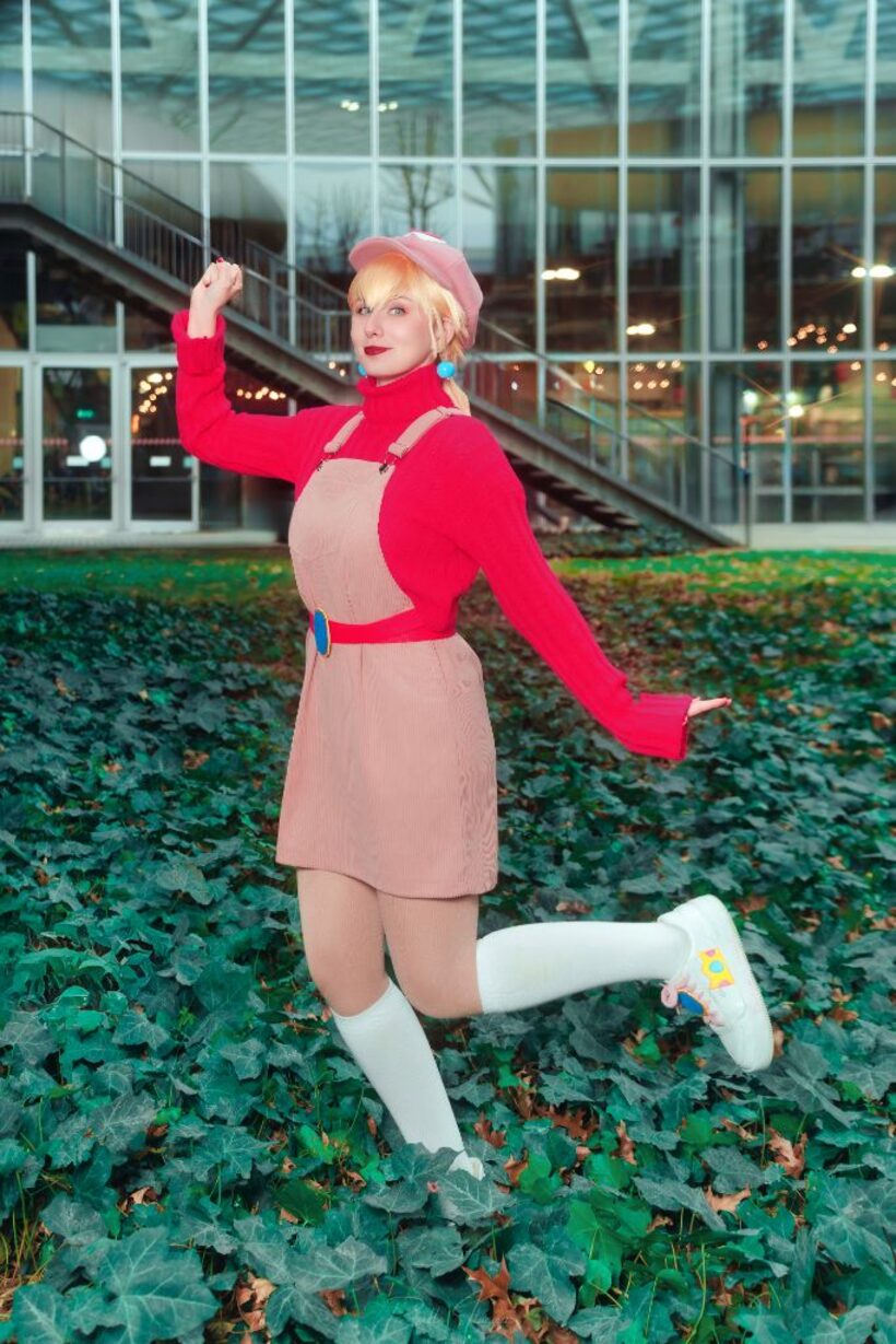 Cosplayer con maglione rosso e basco rosa in posa dinamica su un tappeto di edera